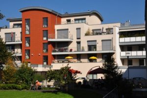 Ein modernes Mehrfamilienhaus mit cremefarbenen und roten Außenwänden, mehreren Balkonen mit gelben Sonnenschirmen und einem gepflegten grünen Rasen mit Bäumen und Sitzgelegenheiten im Freien im Vordergrund.