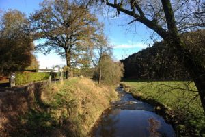 Ein kleiner Bach fließt zwischen grasbewachsenen Ufern und Bäumen unter einem blauen Himmel. Links befindet sich eine Straße mit einem Zaun und Verkehrsschildern. Auf der rechten Seite befindet sich ein offenes grünes Feld, das von bewaldeten Hügeln begrenzt wird.