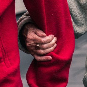 Nahaufnahme einer älteren Hand, die einen Ring trägt und sanft den Arm einer anderen Person in einem roten Mantel hält, was auf Fürsorge und Unterstützung hindeutet.