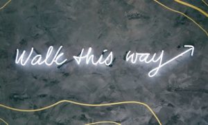 Ein kursives Neonschild mit der Aufschrift "Walk this way" und einem Pfeil, der nach rechts zeigt, auf einer grauen Wand mit gelben, geschwungenen Linien.