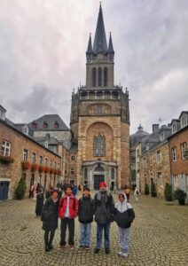 Fünf Menschen stehen lächelnd vor einer historischen Kirche mit einem hohen Kirchturm in einem kopfsteingepflasterten Innenhof, umgeben von alten Backsteingebäuden unter einem bewölkten Himmel.