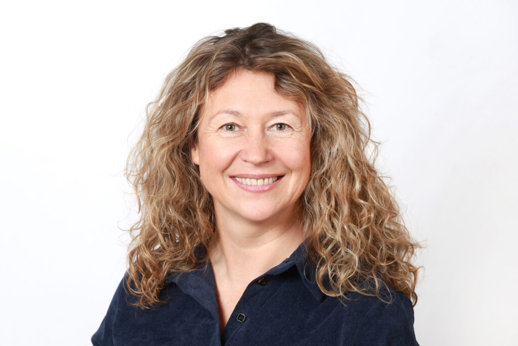 Eine Frau mit gewelltem, hellbraunem Haar und blauem Hemd lächelt in die Kamera vor einem schlichten weißen Hintergrund.